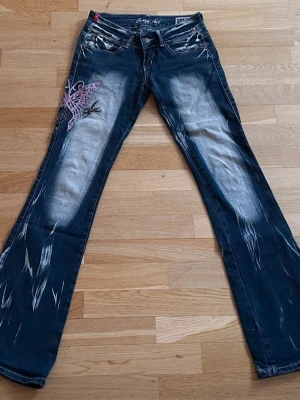 Crazy age blå bootcut jeans med fjärilsbrodyr extremt low waist W26 - Skicka gärna bud!! Säljer ett par blå bootcut jeans från Crazy Age med slitningar och en stor lila fjärilsbrodyr på vänster lår. Jeansen har låg midja, fem fickor och kontrastsömmar. Materialet är klassisk denim med stretch för extra komfort.