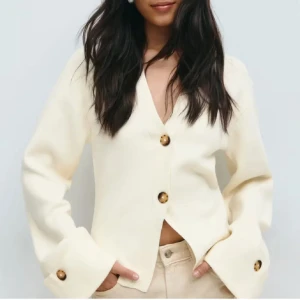 Vit kofta  - Snygg beige kofta med v-ringning och stora bruna knappar framtill och vid ärmsluten. Koftan har långärmade, lite vidare ärmar 