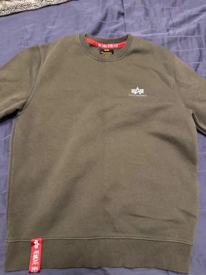 Olivgrön sweatshirt från Alpha Industries - Cool olivgrön sweatshirt från Alpha Industries med logga på bröstet och en röd detalj med texten 'Remove Before Flight' vid nederkanten. Tröjan har rund hals och är tillverkad i mjukt material som känns riktigt skönt. Perfekt för dig som gillar streetwear och militärstil.