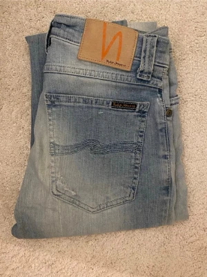 Ljusblå  jeans från Nudie Jeans - Säljer ett par ljusblå skinny jeans från Nudie Jeans med klassisk femficksmodell och slitna detaljer. Jeansen har en snygg tvätt med ljusa partier och Nudie-logga på bakfickan samt läderpatch i midjan. Tillverkade i bomull för skön passform.