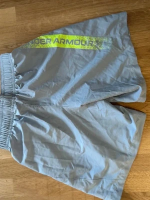 Ljusgrå träningsshorts från Under Armour - Säljer ett par ljusgrå träningsshorts från Under Armour med elastisk midja och sidofickor. Shortsen har en stor logga i neongult på sidan och är tillverkade i ett lätt och snabbtorkande syntetmaterial.