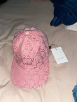 Rosa Gucci keps med GG-mönster - Säljer en rosa keps från Gucci med det klassiska GG-monogrammet över hela kepsen. Kepsen har böjd skärm, justerbart band bak och är tillverkad i canvasmaterial. Perfekt för dig som vill ha en trendig accessoar med lyxig känsla.