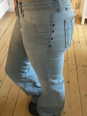 Ultra lowrise bootcut jeans  med knappar - Säljer ett par jättefina ljusblå bootcut jeans med dekorativa knappar längs fickan och klassisk femficksdesign. Jeansen har en väldigt låg midja. 