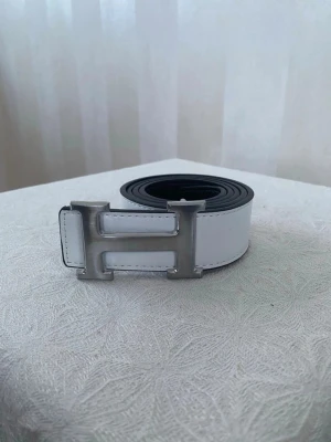 Vitt skärp med H-spänne - Snyggt vitt skärp med ett stort H-format spänne i silverfärg. Bältet har en stilren design och är tillverkat i skinn. Perfekt accessoar för att lyfta din outfit med en exklusiv touch.