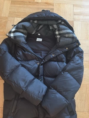 Svart pufferjacka från Burberry - Snygg svart pufferjacka från Burberry med stor huva och klassiskt rutigt foder. Jackan har dragkedja och knappar framtill samt två sidofickor med dragkedja. Perfekt för kalla vinterdagar och riktigt stilren look. Och de står Burberry på varenda zip och knapp som finns på jackan.