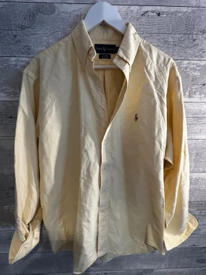 Gul skjorta från Ralph Lauren - Klassisk beige skjorta från Ralph Lauren med knappstängning och broderad logga på bröstet. Skjortan har lång ärm, krage och är tillverkad i mjuk bomull. Perfekt för en clean och stilren look.