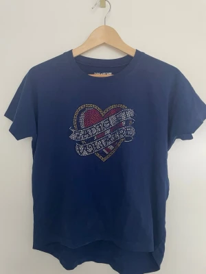 Zadig&voltaire tröja - Mörkblå t-shirt från Zadig&voltaire med ett stort hjärtmotiv framtill, dekorerat med färgglada nitar i svart, rött, gult och vitt. Texten på hjärtat är gjord av små glittriga stenar. Klassisk rund hals och korta ärmar.