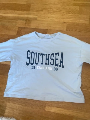 Ljusblå långärmad Southsea tröja - Ljusblå långärmad tröja från Mango med tryck i mörkblått och vitt framtill där det står 'SOUTHSEA 19 ENGLAND 96'. Skön och avslappnad passform, perfekt till jeans eller shorts. Tillverkad i mjuk bomull. Minsta lilla fläck i underarmen som inte syns!! Se bild 4.