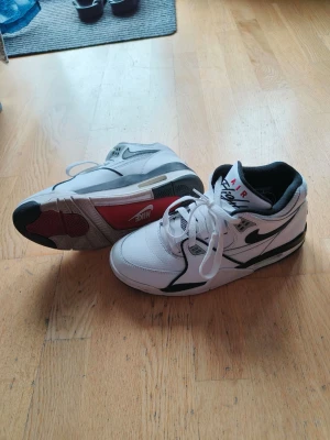 Nike Air Flight vita sneakers - Nike Air Flight sneakers i vitt läder med svarta detaljer och röd logga på plösen. Klassisk retrostil med meshpaneler för ventilation och en chunky sula med svart och röd detalj på sidan. Swoosh-logga på sidan och snörning framtill. Ny pris 1599 kr