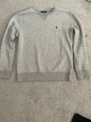 Grå sweatshirt från Polo Ralph Lauren - Klassisk ljusgrå sweatshirt från Polo Ralph Lauren med rund halsringning och diskret broderad logga på bröstet. Tröjan har ribbade muddar vid ärmslut och nederkant, perfekt för en clean och avslappnad stil. Storleken är 14-16 år så uteslog att den är runt xs. Pris kan diskuteras till viss del. Hör av er vid frågor!!