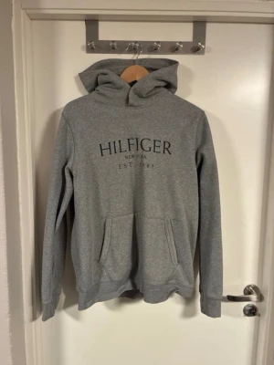 Tommy hilfiger Hoodie  - Snygg grå hoodie från Hilfiger med svart tryck på bröstet där det står 'HILFIGER NEW YORK EST 1985'. Klassisk känguruficka framtill och stor huva. Tillverkad i mjukt sweatshirtmaterial, perfekt för en avslappnad stil.