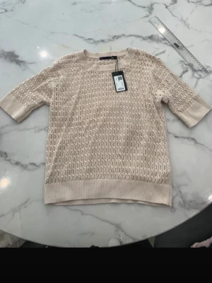 Beige stickad topp med kort ärm - Snygg beige stickad topp med korta ärmar och rund hals. Tröjan har ett diskret hålmönster över hela plagget och bred ribbad kant nedtill. Perfekt för lager på lager och enkel att matcha till olika outfits.