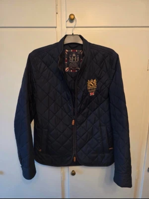 Morris quiltad jacka marinblå herr - Snygg quiltad jacka från Morris i marinblått med guldbroderad logga och brittisk flagga på bröstet. Jackan har ståkrage i manchestertyg, dragkedja och två fickor framtill. Perfekt för dig som gillar klassisk och stilren look.