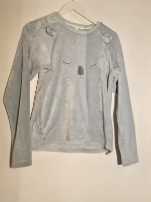 Ljusblå loungeweartröja med koala - Supermjuk ljusblå loungeweartröja i polyester broderat koalaansikte och små öron på axlarna. Rund hals och långa ärmar. Perfekt för myskvällar eller chill hemma. Materialet är fluffigt och känns riktigt skönt mot huden.