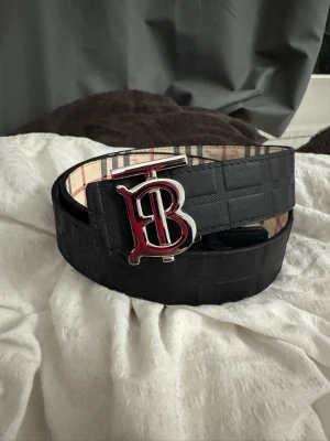 Svart Burberry bälte med rutigt foder - Snyggt svart bälte från Burberry med klassiskt rutigt foder på insidan och en stor metallspänne med Burberrys logotyp. Bältet har en stilren design och passar perfekt till jeans eller kostymbyxor.