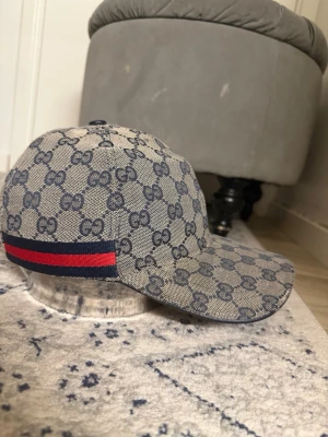 Gucci keps med monogram och röd-blå rand - Snygg keps från Gucci i beige med mörkblått GG-monogram över hela kepsen. Klassisk böjd skärm och en ikonisk röd-blå rand på sidan. Tillverkad i canvasmaterial som ger en exklusiv känsla och streetstyle-vibe.