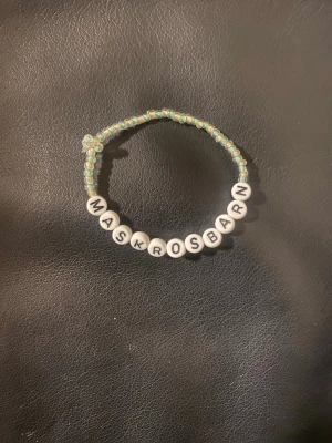 Pärlarmband med texten MASKROSBARN - Handgjort armband med små gröna och vita pärlor samt vita bokstavspärlor som stavar 'MASKROSBARN'. Elastisk tråd gör det enkelt att ta på och av. Perfekt accessoar för dig som vill bära ett personligt budskap.
