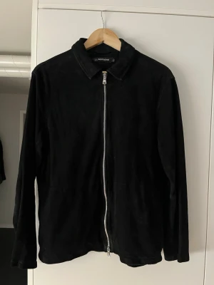 Svart mockajacka från Lindeberg - Stilren svart mockajacka från Lindeberg med rak passform och klassisk krage. Jackan har en silverfärgad dragkedja framtill och långa ärmar. Perfekt för dig som gillar minimalistisk och tidlös stil. Liten i storleken sitter som S. Kvitto finns!