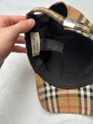 Burberry beige rutmönstrad keps - Säljer en klassisk keps från Burberry i det ikoniska beige rutmönstret med svarta, vita och röda detaljer. Kepsen har justerbart spänne i metall baktill och är fodrad med svart tyg på insidan. Perfekt accessoar för en trendig och tidlös look.