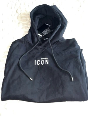 Svart hoodie från Dsquared2 ICON - Svart hoodie från Dsquared2 med vit text 'ICON' på bröstet och justerbar huva med snören. Klassisk passform och mjukt material, perfekt för en avslappnad streetwear-look. Snygg och enkel design som funkar till det mesta.