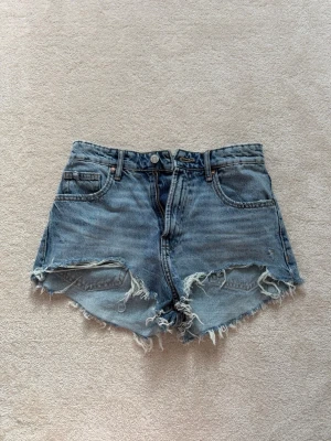 Blå jeansshorts från Zara - Säljer ett par blå denimshorts med hög midja och råa, fransiga kanter. Klassisk femficksmodell med dragkedja och knapp framtill. Perfekta för sommaren och ger en avslappnad vibe.