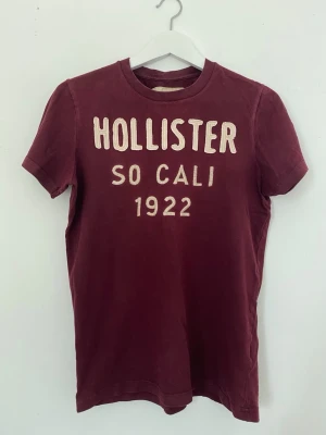 VIntage vinröd t-shirt från Hollister - Snygg vinröd t-shirt från Hollister med tryck 'HOLLISTER SO CALI 1922' på bröstet. Klassisk passform med rund hals och korta ärmar. Tillverkad i mjuk bomull som känns skön mot huden. Perfekt för en avslappnad och trendig look. Köpt tidigt 2000-tal.