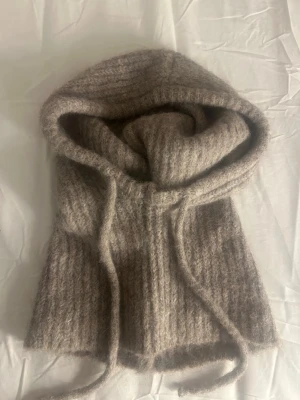 Beige stickad balaklava med snören - Mysig beige stickad balaklava med dragsnören framtill. Tillverkad i ett mjukt, fluffigt material som ger en avslappnad look. Perfekt för att hålla värmen och addera stil till din outfit.