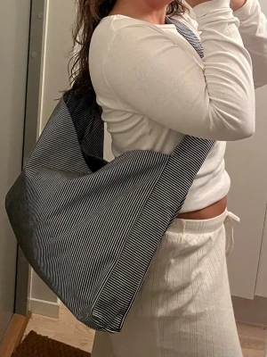 Randig svart axelväska i tyg - Säljer en stor axelväska i blått tyg med tunna vita ränder. Väskan har en bred axelrem och rymlig form, perfekt för att bära med sig allt man behöver. Enkel och stilren design som passar till många outfits.