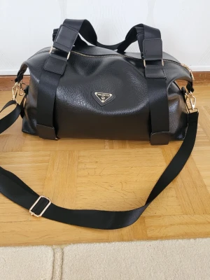 Svart weekendbag från Walentia - Snygg svart handväska från Walentia i konstläder med guldfärgade detaljer. Väskan har justerbar axelrem, två breda handtag och en dragkedja upptill.  Köptes inte länge sen amvönd en gång på resa men råkade få en liten skav på botten. Se bild.  Längd 44cm höjd 30cm bredd 22cm