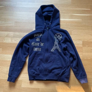 Bling licid hoodie (Mörkblå) - Snygg mörkblå hoodie med dragkedja och paljettdetaljer i form av text och Eiffeltornet. Perfekt för en avslappnad stil med en touch av Paris. Den har långa ärmar och en praktisk huva med snörning.