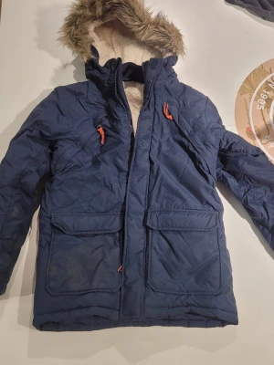 Mörkblå parkas med fuskpäls - Snygg mörkblå parkas med huva och fuskpäls, röda detaljer på dragkedjorna och två stora fickor framtill. Jackan har ett badge på ärmen med bergsmotiv och texten Mount Hood Oregon State. Fodrad med mjukt material för extra värme.