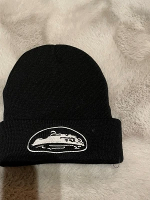 Svart ribbstickad mössa med brodyr - Säljer en svart ribbstickad mössa med uppvikt kant och en broderad vit patch framtill som föreställer ett fordon, troligen en ubåt. Mössan är enkel och stilren, perfekt för kalla dagar och passar till de flesta outfits.