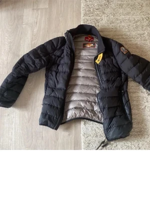 Svart dunjacka från Parajumpers - Snygg svart dunjacka från Parajumpers med klassisk quiltad design och grå insida. Jackan har dragkedja, hög krage och ikonisk patch på ärmen. Perfekt för kalla dagar och riktigt varm tack vare dunfyllningen.  Tyvär trasig på ena axeln men sy ihop och du har en hel jacka! Den är äkta! 