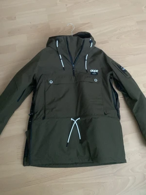 Olivgrön vindjacka från Dope (intressekoll!!) - Snygg olivgrön vindjacka från Dope med huva, halv dragkedja framtill och stor magficka med vita detaljer. Jackan har dragsko i midjan och svarta dragkedjor i sidorna. använt 3 antal gånger och har inga defekter! nyköpt för 2099kr