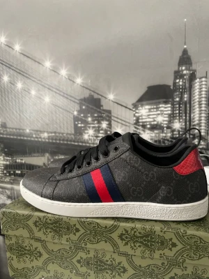 Gucci svarta sneakers med röd/blå detalj - Snygga svarta sneakers från Gucci med diskret GG-mönster i tyget och klassiska röd/blå ränder på sidan. Skon har en vit sula och röda detaljer vid hälen. Materialet är canvas med läderdetaljer och låg profil. Perfekt för dig som vill ha en exklusiv look.