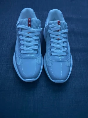 Vita sneakers från Prada - Snygga vita sneakers från Prada med klassisk siluett, meshpaneler och detaljer i blankt läder. Skorna har vita snören och en röd logotyp på plösen. Perfekta för dig som gillar stilrena och exklusiva sneakers.