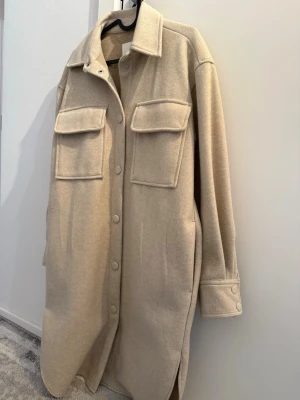 Beige overshirt med stora fickor - Stilren beige overshirt i mjukt material med två stora bröstfickor och knappar framtill. Lång modell med klassisk krage och knappdetaljer vid ärmsluten. Perfekt lager-på-lager-plagg för en trendig look.