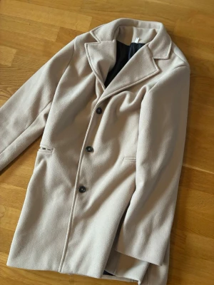 Beige kappa med knappar - Stilren beige kappa med tre svarta knappar framtill och klassisk krage. Kappan har två fickor och är fodrad med svart tyg på insidan. Perfekt för dig som vill ha en enkel men snygg look till hösten eller vintern.