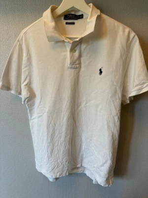 Vit pikétröja från Polo Ralph Lauren - Klassisk vit pikétröja från Polo Ralph Lauren. Tröjan har korta ärmar, krage och knappar framtill samt den ikoniska broderade loggan på bröstet. Tillverkad i mjuk bomull som känns skön mot huden. Skick 9,5/10 använd endast ett fåtal gånger.