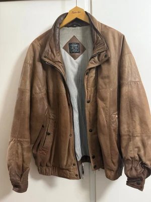 Brun skinnjacka från Donar - Säljer en brun skinnjacka från Donar med klassisk bombermodell. Jackan har dragkedja, tryckknappar, två snedställda fickor fram och ribbade muddar vid ärmslut och midja. Insidan är fodrad med randigt tyg och kragen är hög och kan stängas med knapp.