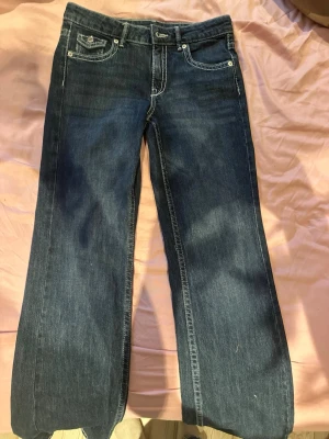 Mörkblå bootcut jeans med kontrastsömmar - Säljer ett par mörkblå bootcut jeans med vita kontrastsömmar och snygga fickdetaljer bak med lock och knapp. Klassisk femficksmodell i mjukt denimtyg, perfekt för en avslappnad men trendig look.