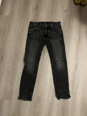 Svarta jeans från Lee - Säljer ett par svarta jeans från Lee med klassisk femficksdesign och diskreta slitningar. Jeansen har normal midja, raka ben och är tillverkade i ett mjukt denimtyg med lite stretch för extra komfort. Perfekta till en avslappnad streetwear-look.