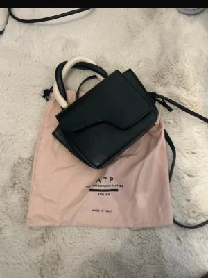 Svart handväska från ATP Atelier - Snygg svart handväska från ATP Atelier med unik asymmetrisk lockdesign och kontrasterande vita handtag. Tillverkad i Italien av slätt läder. Väskan har både handtag och axelrem för olika stilar. Tror inte att färgen på denna väska säljs längre. Har några defekter se bilder 