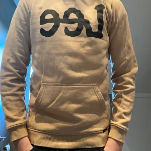 Beige sweatshirt från Lee - Säljer en beige sweatshirt från Lee med stor svart logga på bröstet. Tröjan har lång ärm, ribbade muddar och en stor magficka framtill. Perfekt för en avslappnad och stilren look.