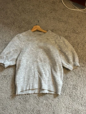 Grå stickad tröja med puffärm  - Mysig grå stickad tröja med korta puffärmar och ribbad kant vid hals, ärmslut och nederkant. Perfekt för lager på lager och enkel att matcha till olika outfits. Tröjan har en rak passform och är gjord i mjukt material.