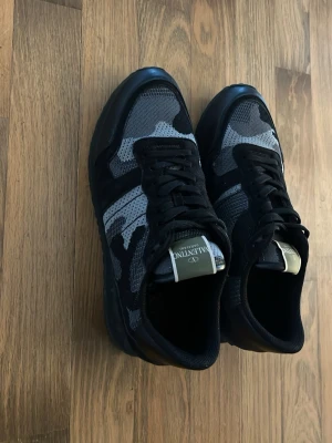 Valentino camo sneakers i svart och grå - Snygga sneakers från Valentino med svart och grått camomönster. Skorna har svarta snören, meshdetaljer och en klassisk låg siluett. Märkeslogga på plösen och robust sula för extra komfort. Perfekta för dig som vill ha en unik och trendig look.