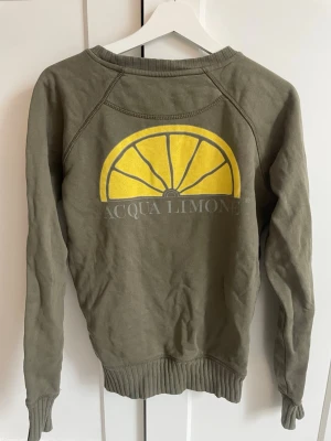 Olivgrön sweatshirt Acqua Limone - Olivgrön sweatshirt från Acqua Limone med klassiskt gult citrontryck och logga på ryggen. Tröjan har rund halsringning, långa ärmar och ribbade muddar vid ärmslut och nederkant. Perfekt för en avslappnad och trendig stil.