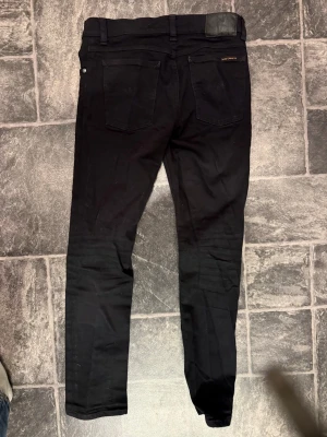 Svarta slim fit jeans från Nudie Jeans.W28 L32 - W28 L 32 . Säljer ett par svarta slim fit jeans från Nudie Jeans med klassisk femficksdesign och läderpatch bak i midjan. Jeansen har smal passform och är tillverkade i stretchigt material för extra komfort. Perfekta för en clean och stilren look. Knappt använda 