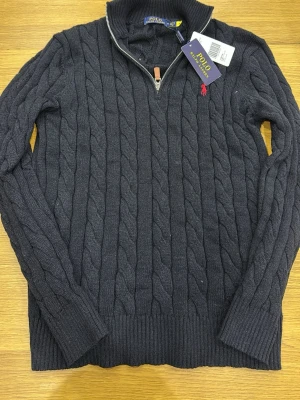 Svart kabelstickad tröja Polo Ralph Lauren - Kabelstickad svart tröja från Polo Ralph Lauren med halv dragkedja och läderdetalj på zippen. Tröjan har lång ärm, ribbade muddar och en röd broderad logga på bröstet. Perfekt för dig som gillar klassisk och stilren look. OBS! fick som present. PRIS KAN DISKUTERAS