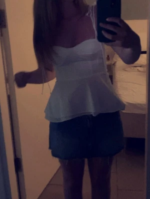 Vit peplum korsettopp med smala band - Supersöt vit korsettopp med smala axelband och peplumvolang. Toppen har markerade kupor och figurnära passform som ger en snygg siluett. Perfekt att styla med jeans eller kjol för en trendig look.